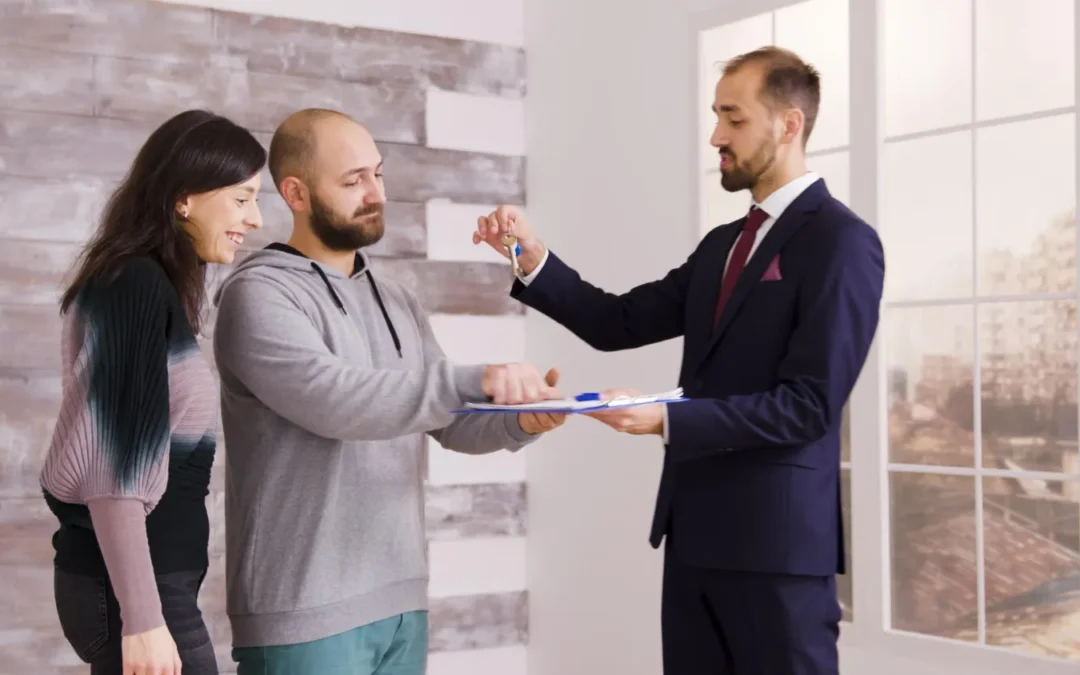 A real estate agent in a suit handing house keys to a young couple while holding a clipboard
